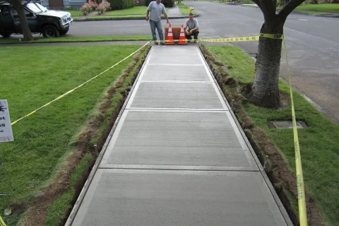 Freshly poured concrete walkway bordered by grass with caution tape and construction materials