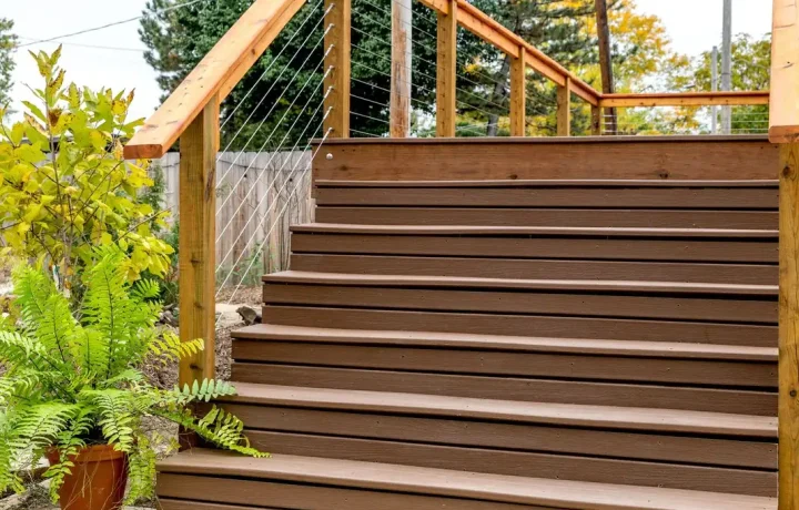 Brown wooden stairs with wire mesh handrails leading up to a porch, surrounded by green plants and shrubs