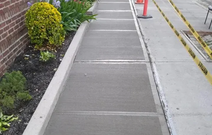 Freshly laid concrete sidewalk with expansion joints, adjacent to a garden bed and brick wall.